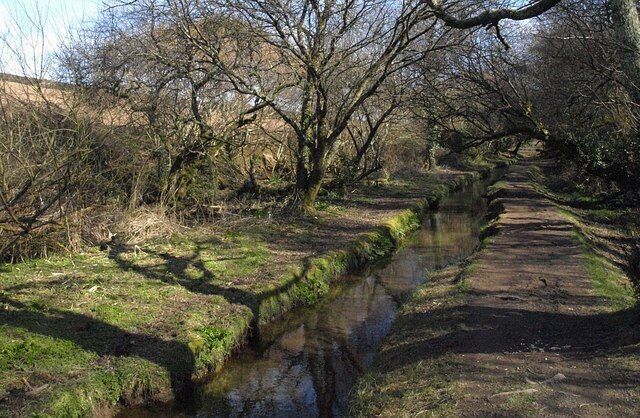 A narrow water channel with a stone path alongside, surrounded by trees and grassy banks.