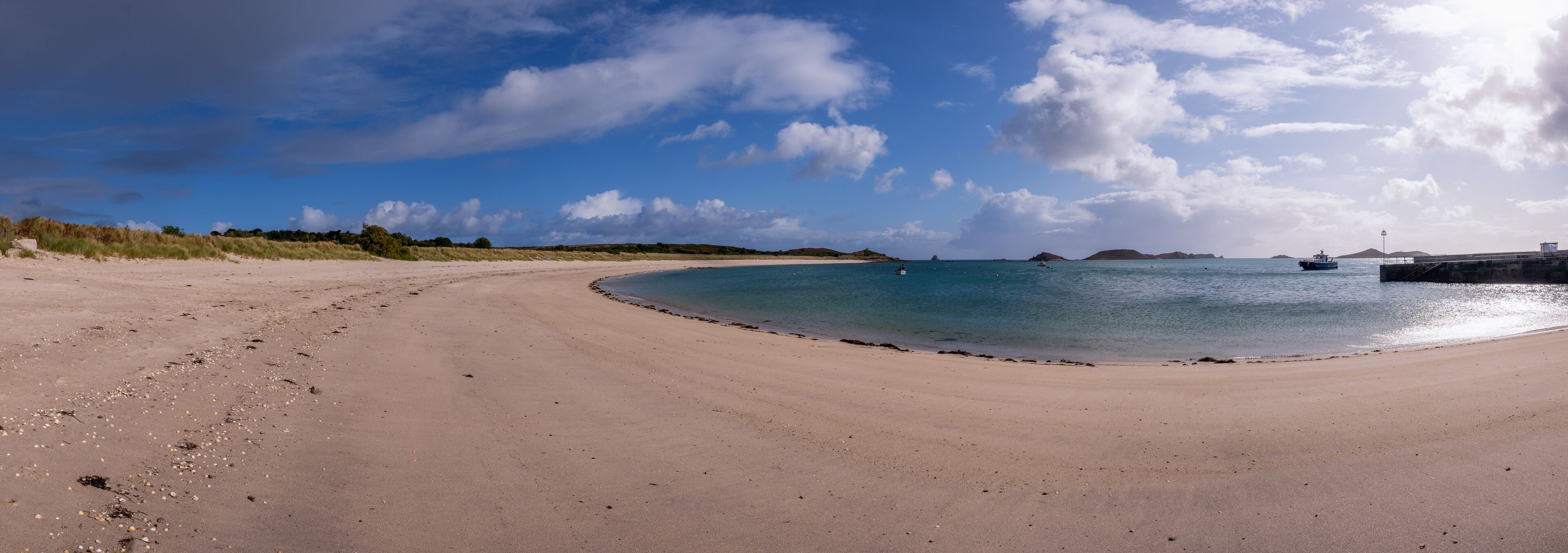 A sandy beach with calm waters, a small boat, and distant islands under a partly cloudy sky.
