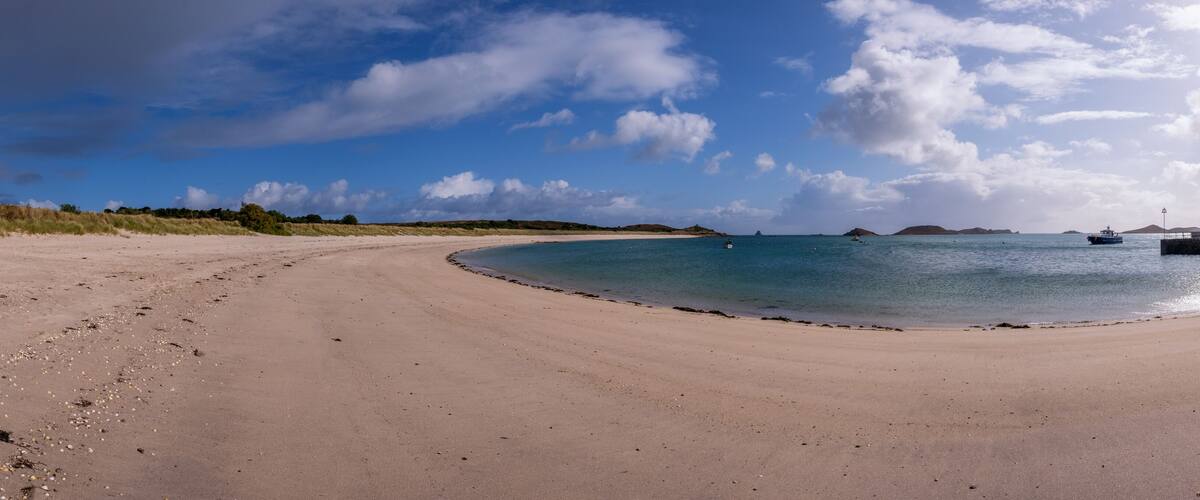 A sandy beach with calm waters, a small boat, and distant islands under a partly cloudy sky.
