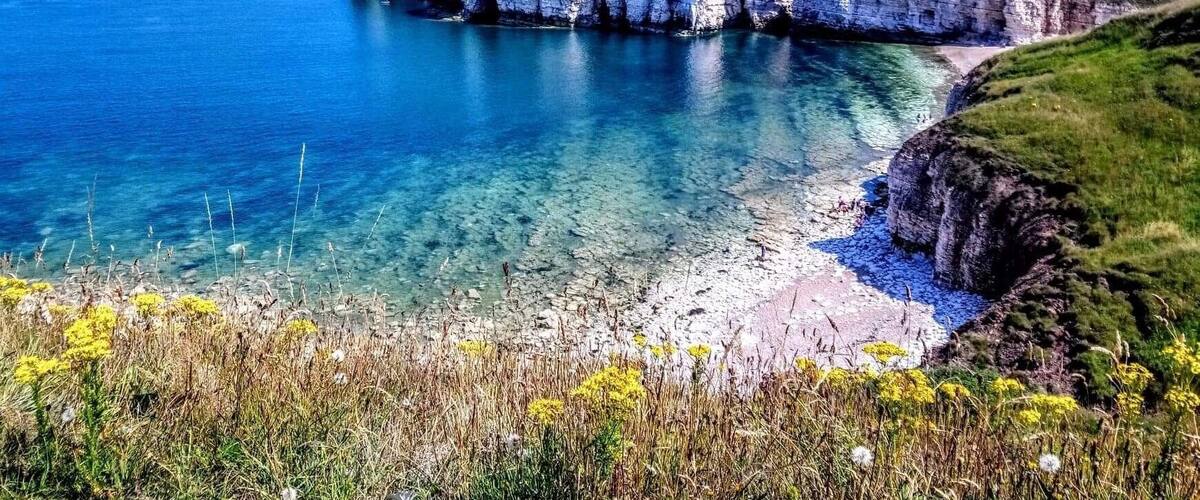 Une falaise côtière surplombant une eau d’un bleu limpide, une plage de sable et des maisons situées en haut.