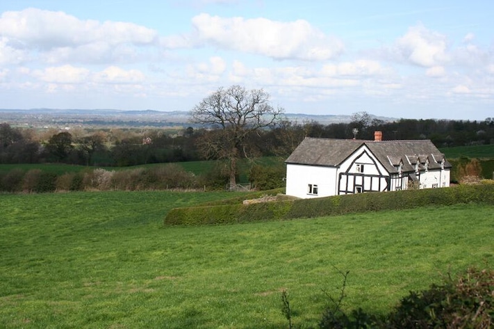 Casa blanca con detalles negros en un entorno rural, rodeada de campos verdes y un árbol.
