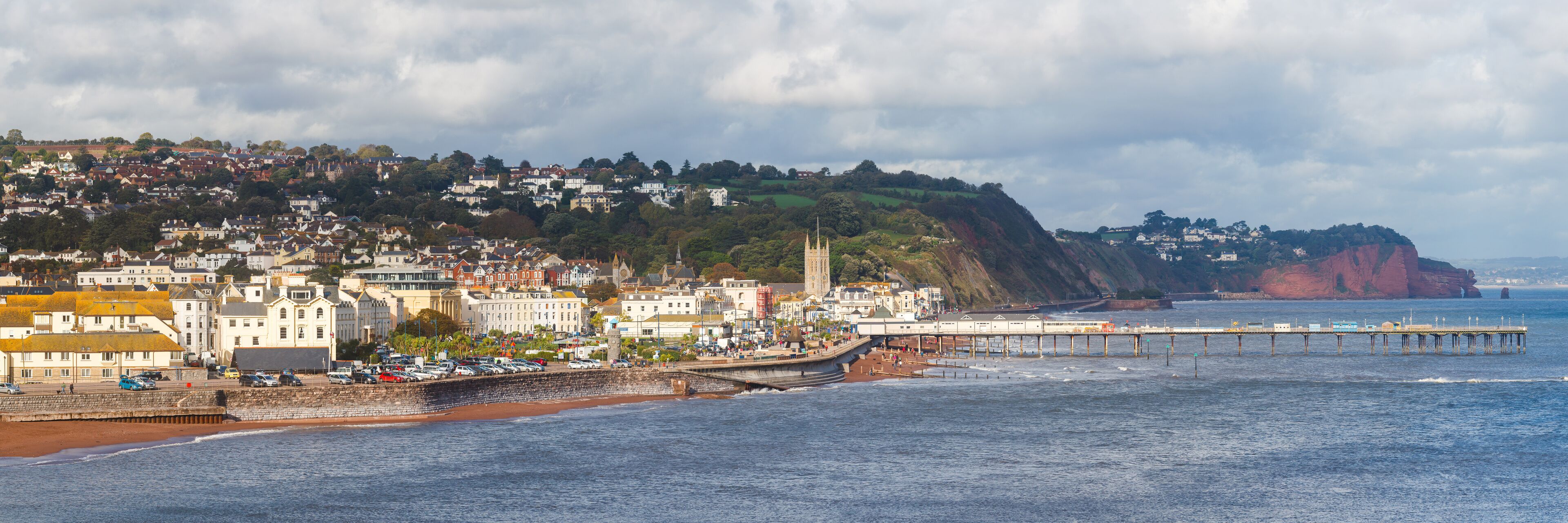 A coastal town with a pier extending into the sea, a prominent church, and a hill with houses.