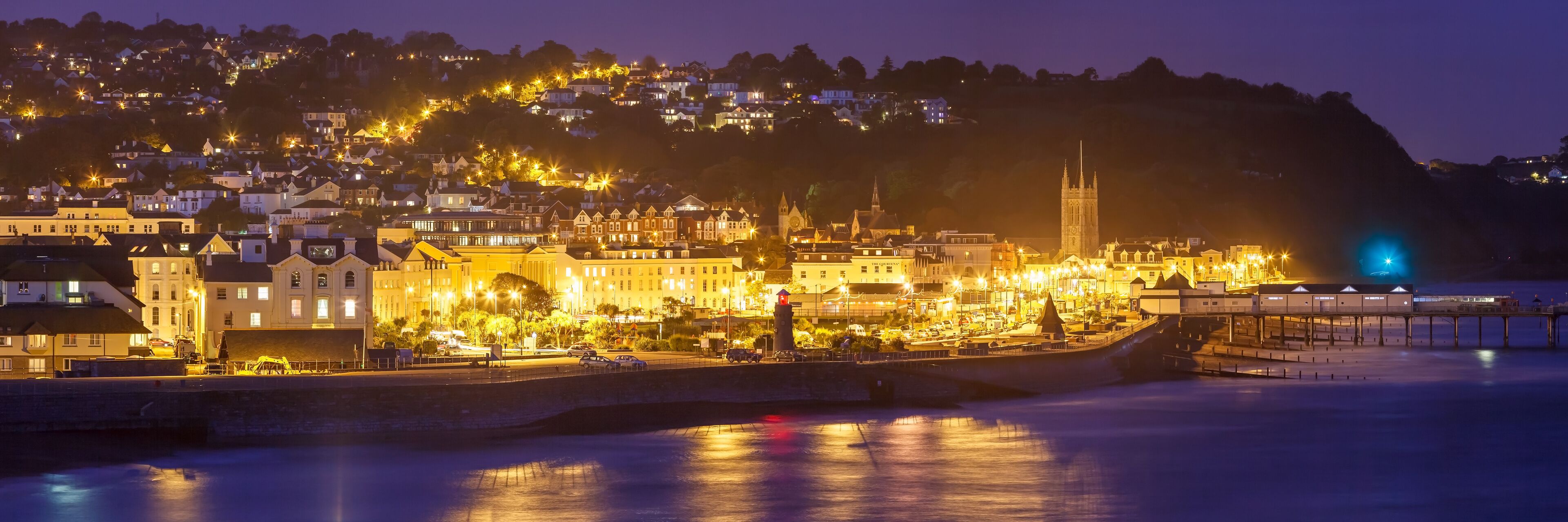 A coastal town at dusk with illuminated buildings, a prominent church tower, and a pier extending into the water.