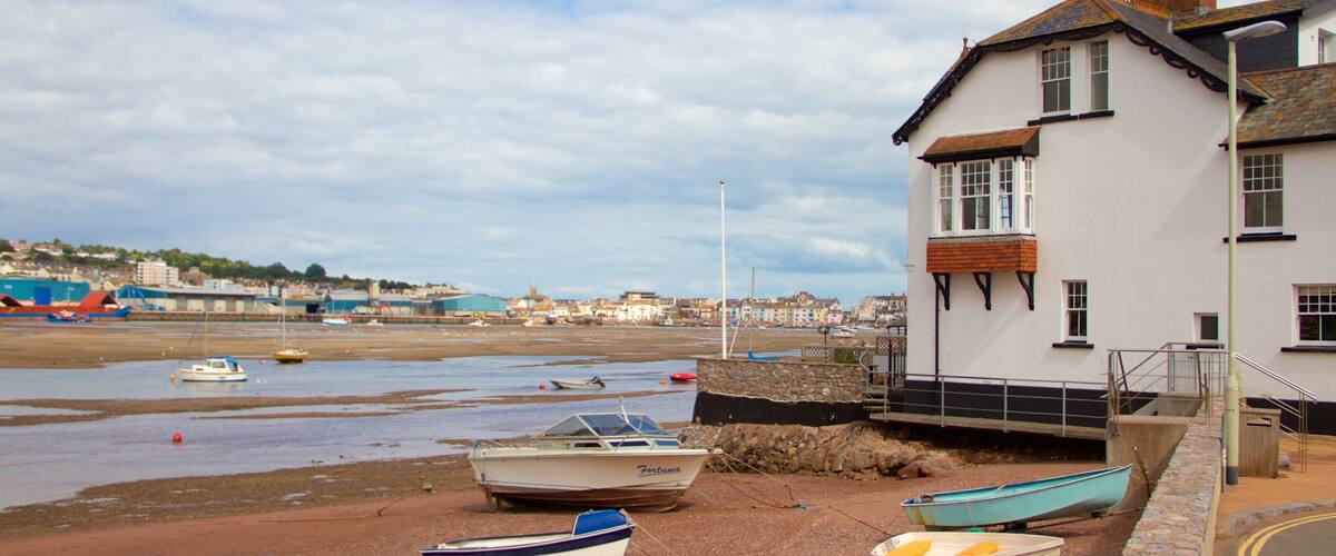 A coastal scene with several small boats tied up on a pebbly beach, a white building with a stone wall, and a distant view of a town with buildings and boats on the water.
