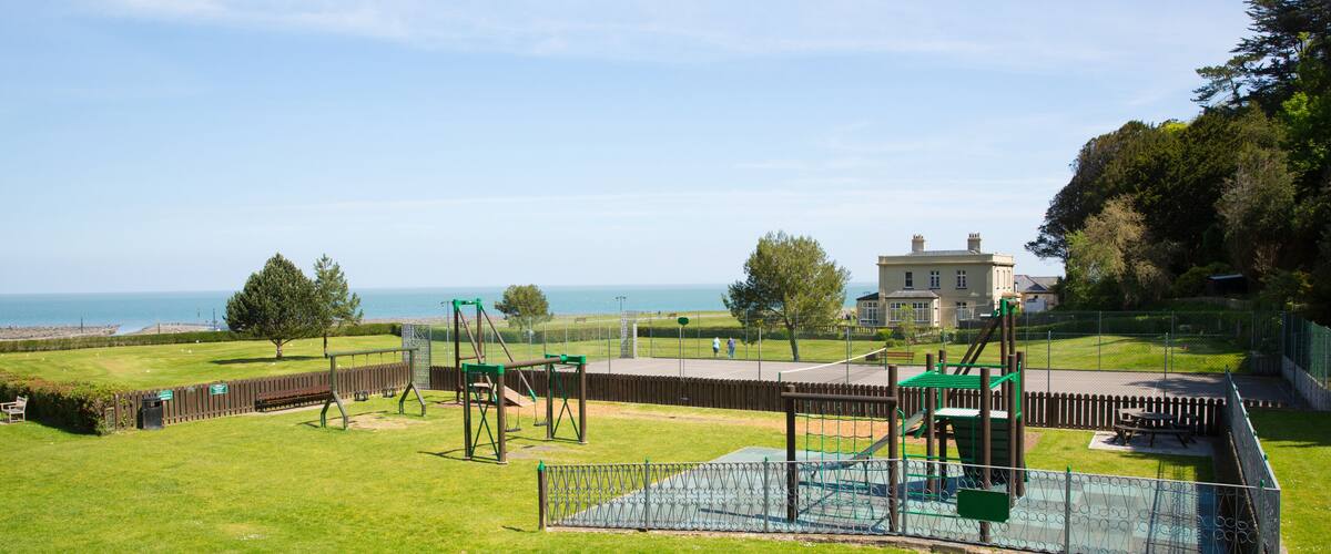 A playground with swings and climbing structures, a fenced area, and a large house with a lawn in the background.