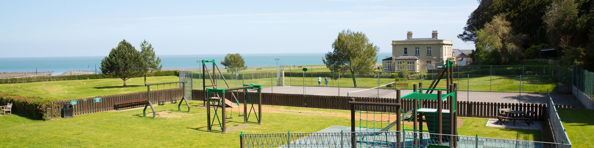 A playground with swings and climbing structures, a fenced area, and a large house with a lawn in the background.