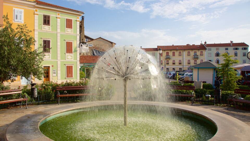 A fountain with water jets in a circular basin, surrounded by colorful buildings and benches.