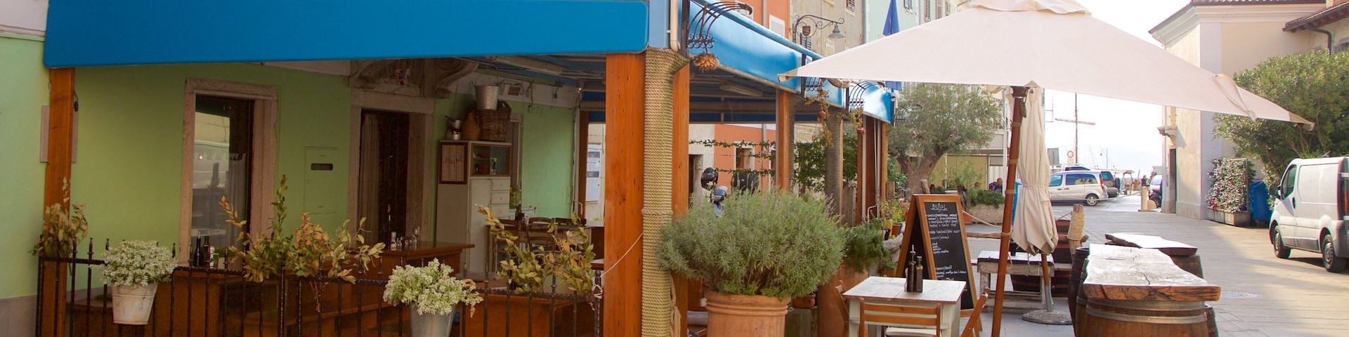 Um café charmoso ao ar livre com mesas e cadeiras de madeira, plantas em vasos e um guarda-sol grande.