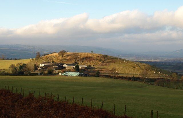 A rural landscape with rolling hills, a fenced field, and a small cluster of buildings.