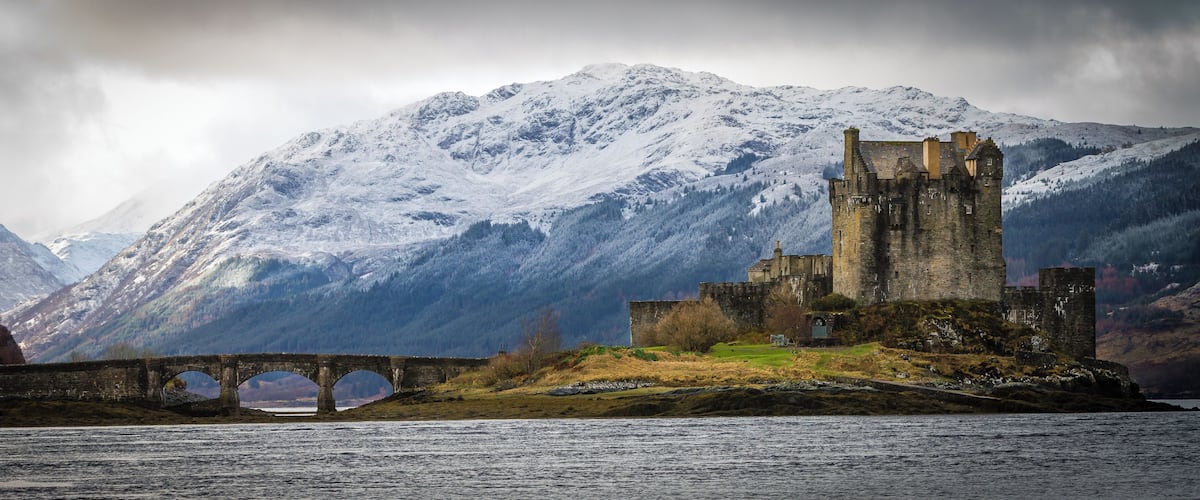 A historic castle on a rocky island with a bridge leading to it, surrounded by snow-covered mountains.