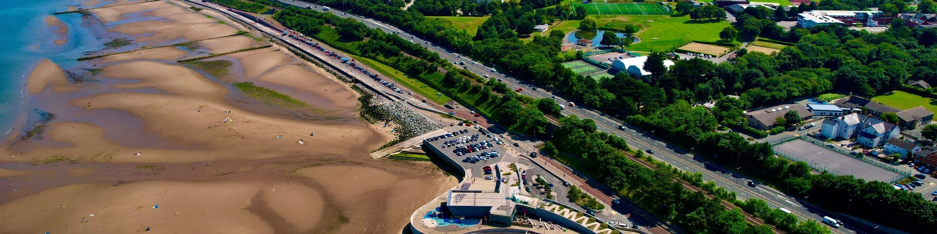 Uma cidade costeira com uma praia, um estacionamento e uma área de parque arborizada.