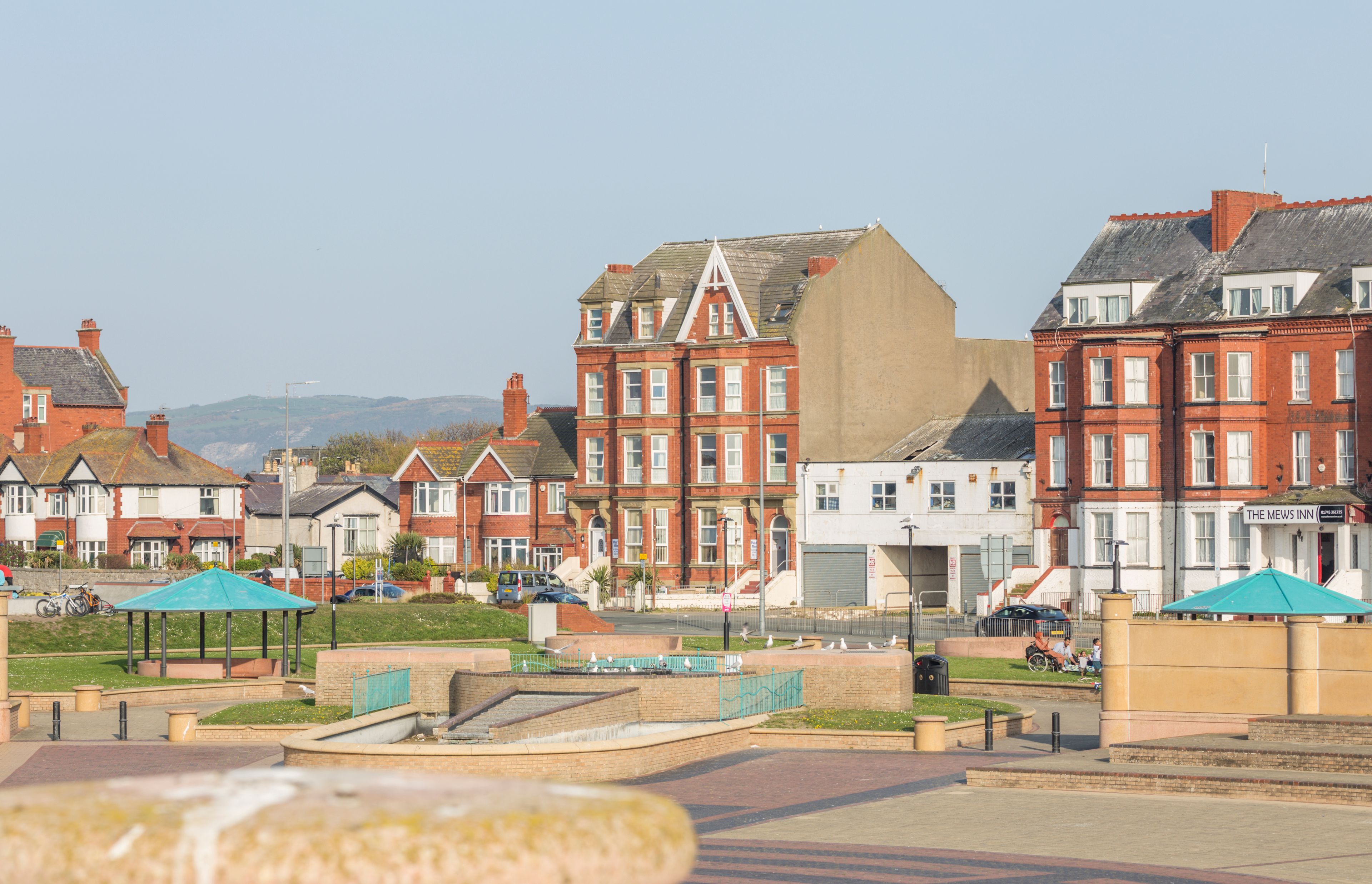 A street with red brick houses, a large building with a flat roof, and a sign for 'The Mews Inn'.