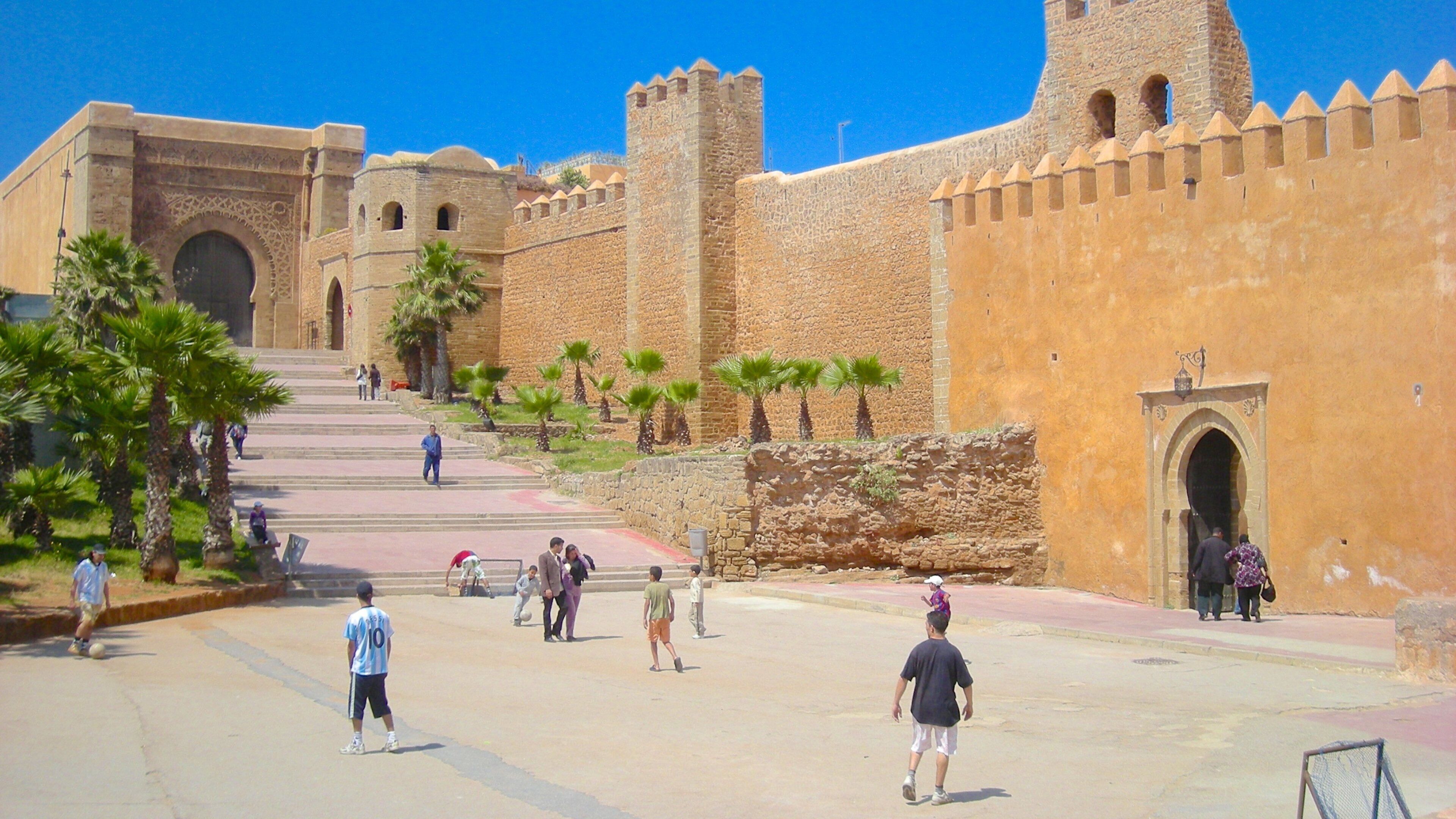 A historic fort with stone walls and towers, palm trees, and people walking around.