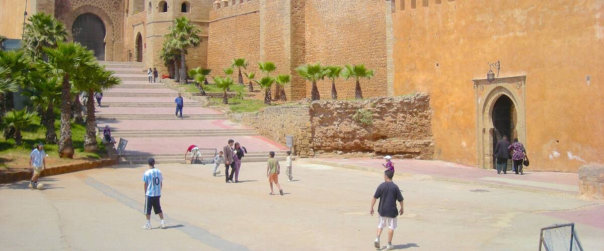 A historic fort with stone walls and towers, palm trees, and people walking around.