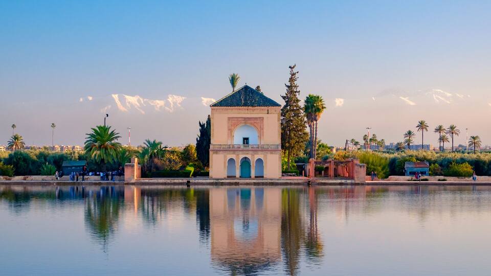 A serene lakeside scene with a historic building featuring an arched entrance, palm trees, and a mountain range in the background.