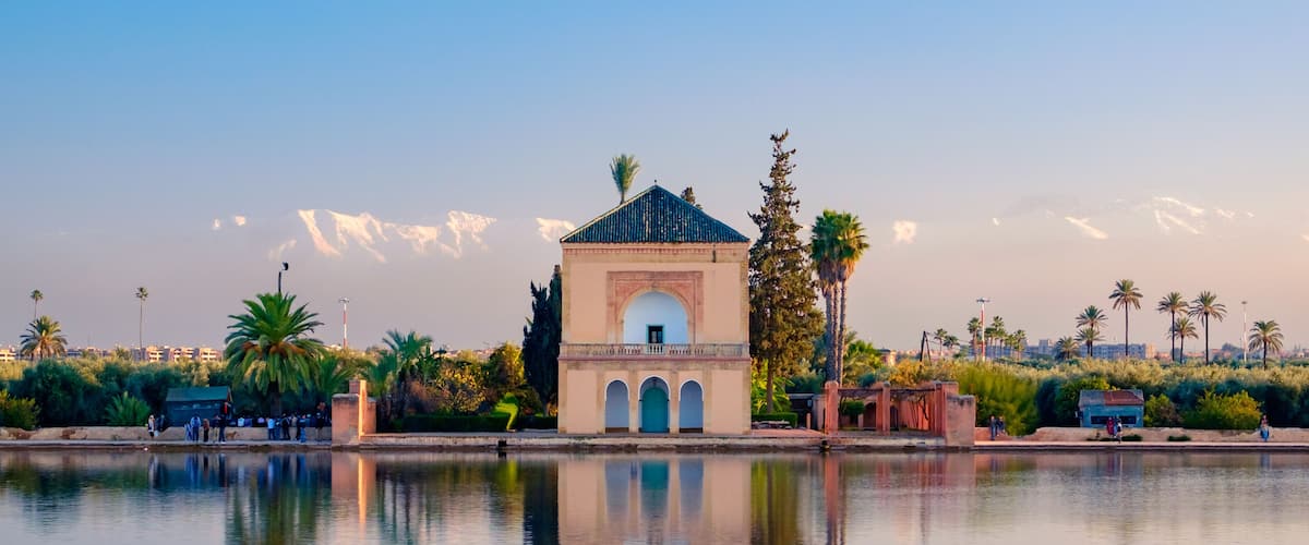 A serene lakeside scene with a historic building featuring an arched entrance, palm trees, and a mountain range in the background.