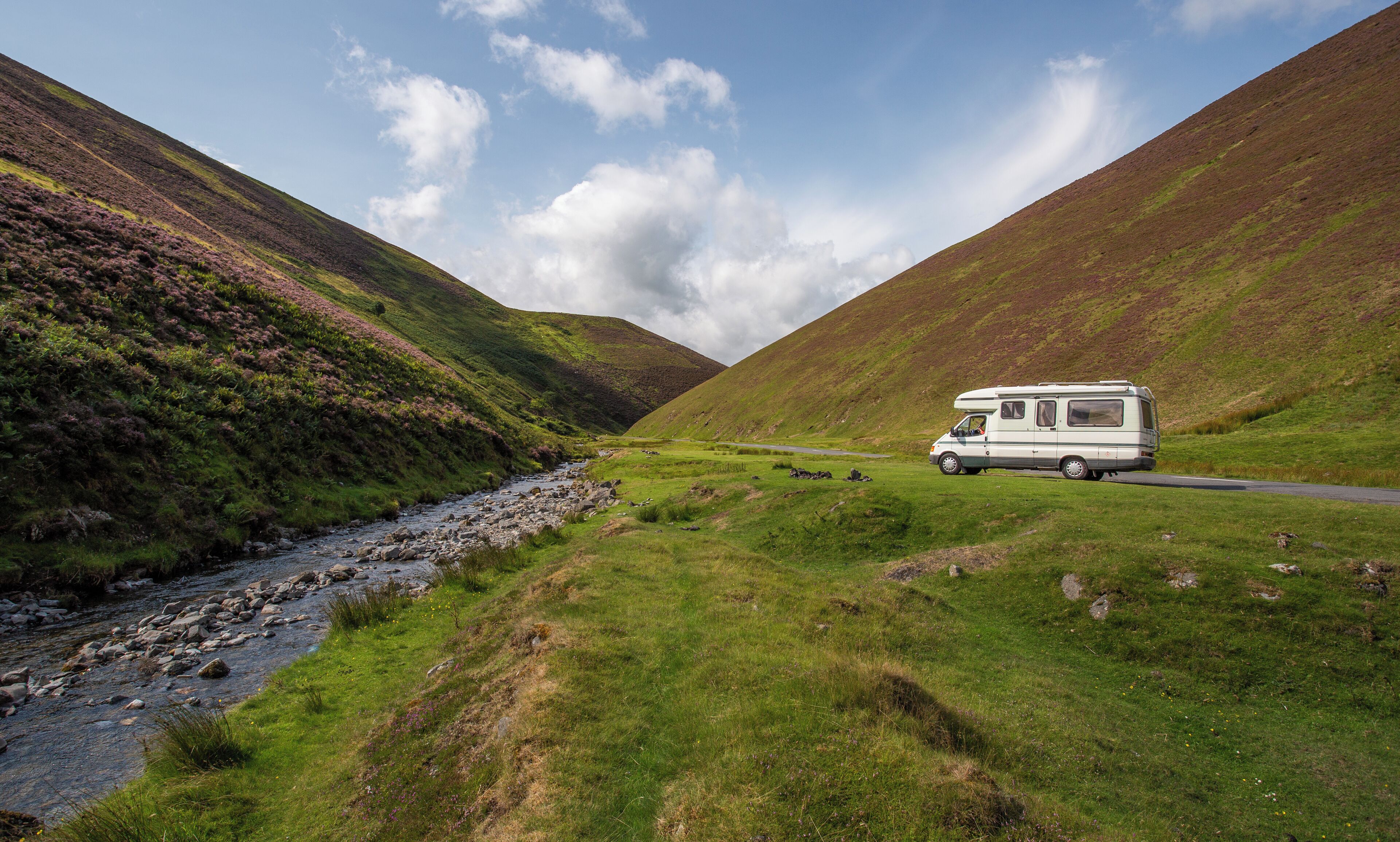 A white camper van parked on a grassy hillside beside a rocky stream.