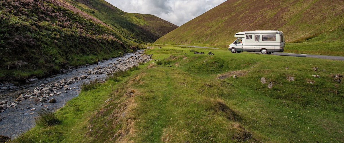 A white camper van parked on a grassy hillside beside a rocky stream.