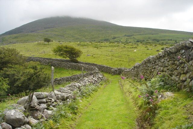 Un muro de piedra, un sendero cubierto de hierba y una colina al fondo.