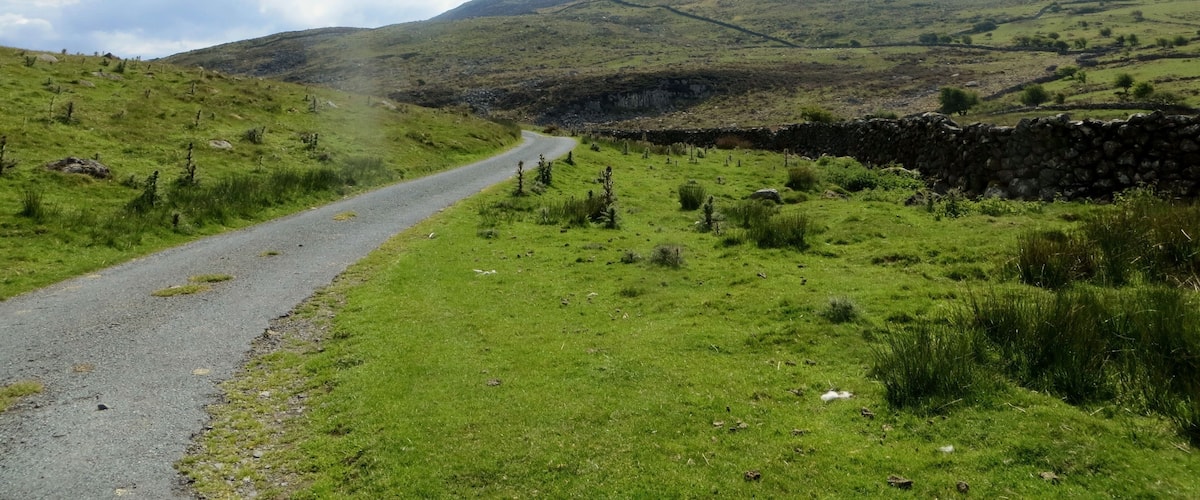A rural road winding through a green landscape with a stone wall and a hill in the background.