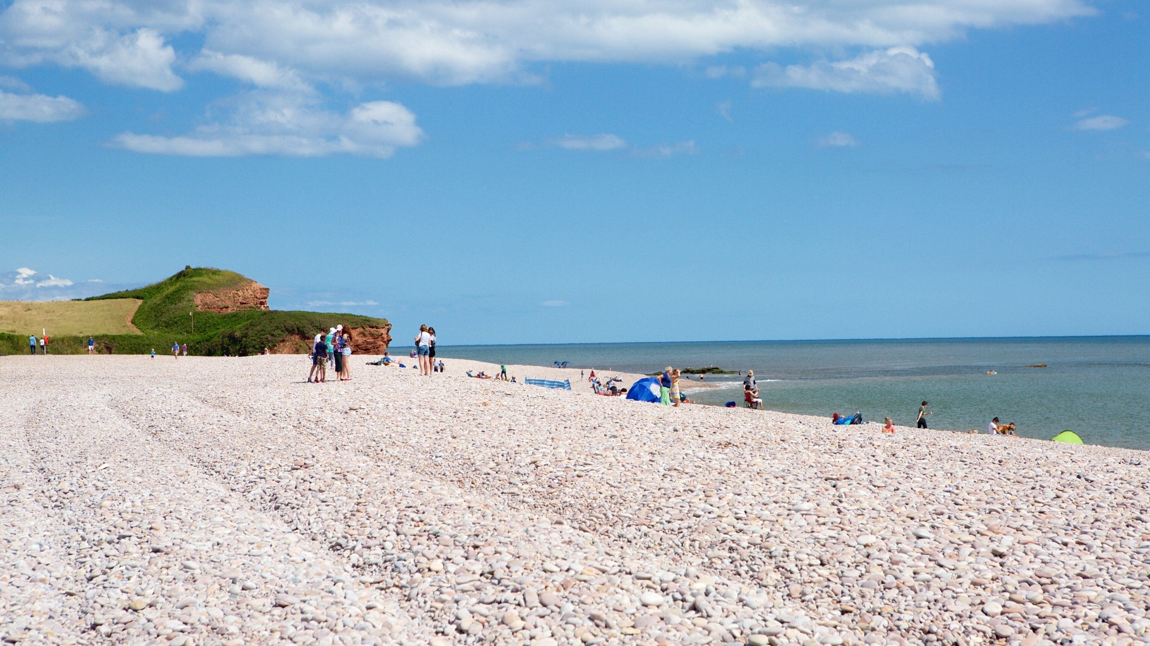 Ein Kiesstrand mit Menschen, ein grasbewachsener Hügel und das Meer.