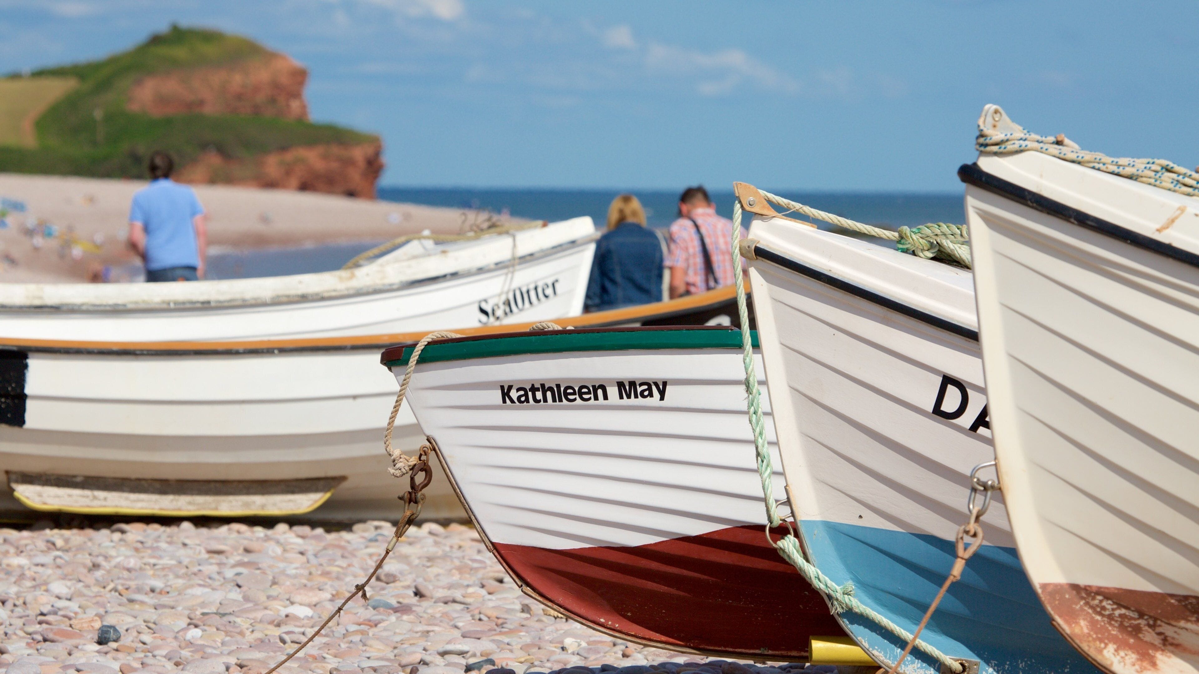 A row of small boats on a pebbled beach with a rocky cliff in the background.