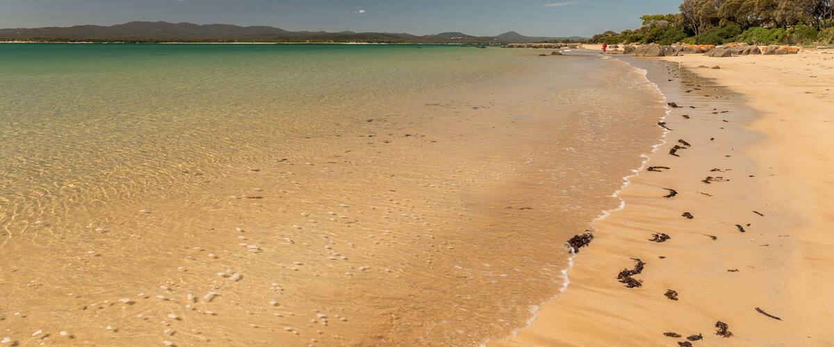 A sandy beach with clear turquoise water, footprints, and distant mountains.