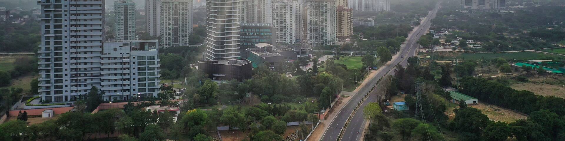 A cityscape with a prominent tower labeled 'Paras Trade Centre' amidst other high-rise buildings, under a cloudy sky with birds flying.