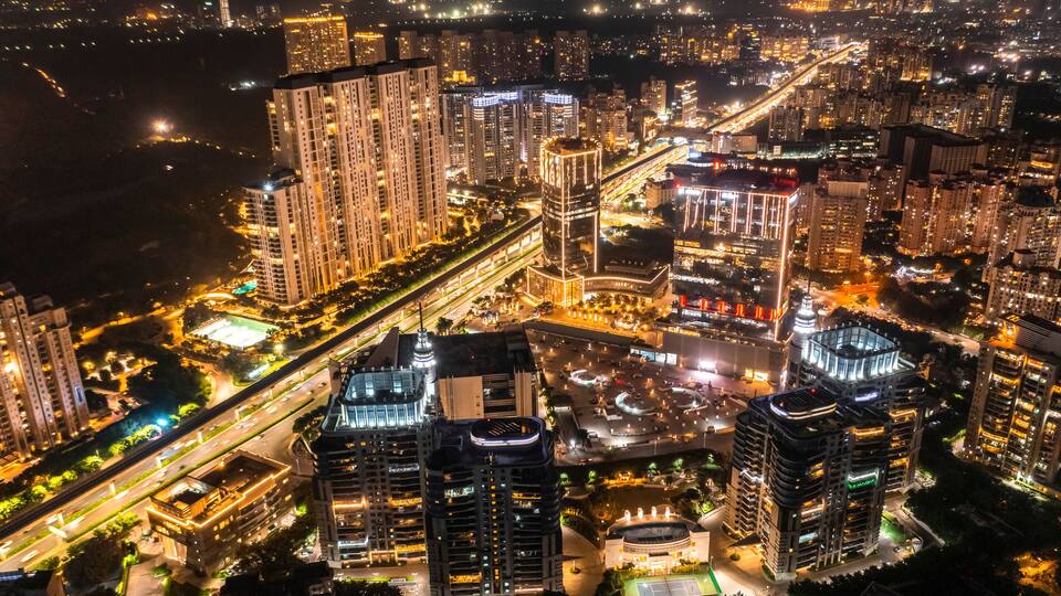 A nighttime aerial view of a densely built urban area with high-rise buildings, illuminated streets, and a prominent roundabout.