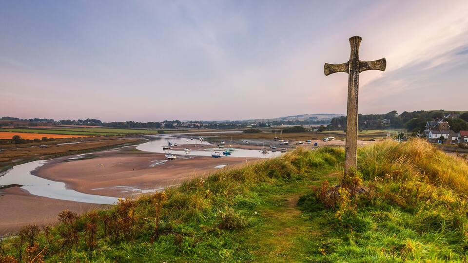 A coastal landscape with a wooden cross, a river, boats, and houses in the background.