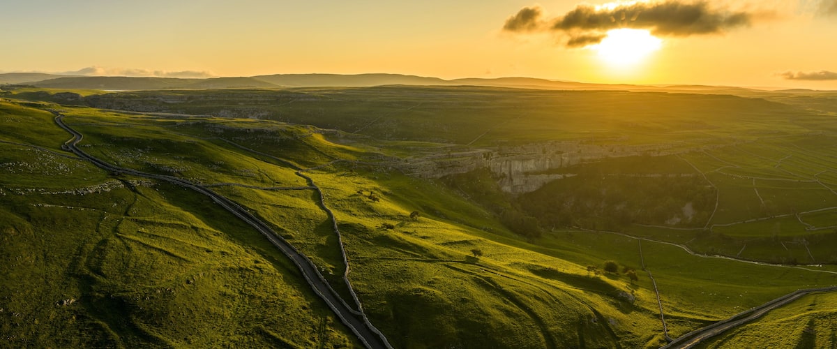 A winding road through rolling green hills with a valley below and the sun setting in the background.