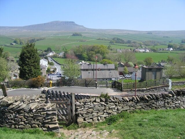 A stone wall, a small building, and a road in a rural area with green fields and a hill in the background.