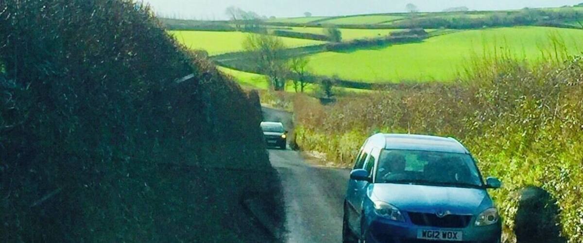 A rural road with a blue car, green fields, and rolling hills in the background.