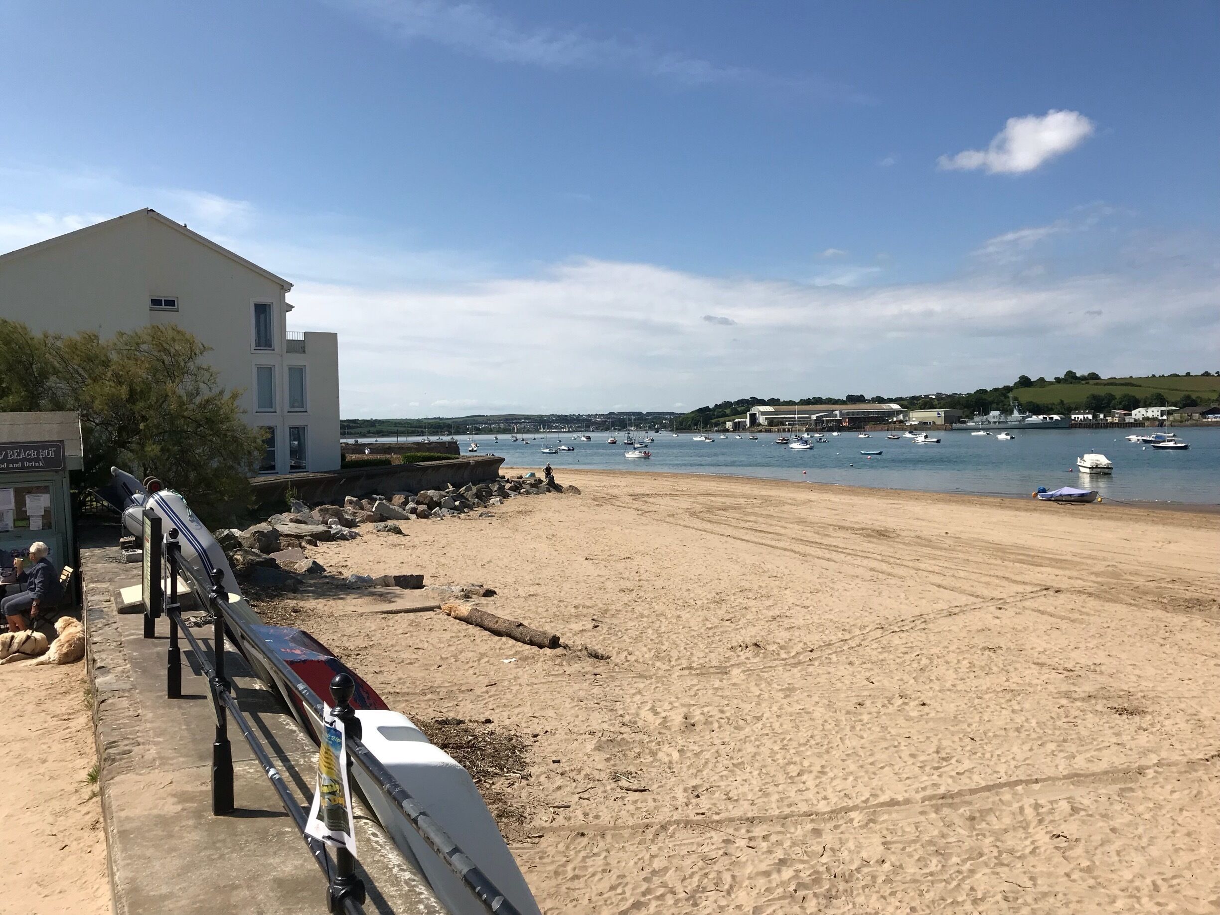 A sandy beach with a white building and a sign that reads "BEACH BAR". Boats are visible on the water in the background.