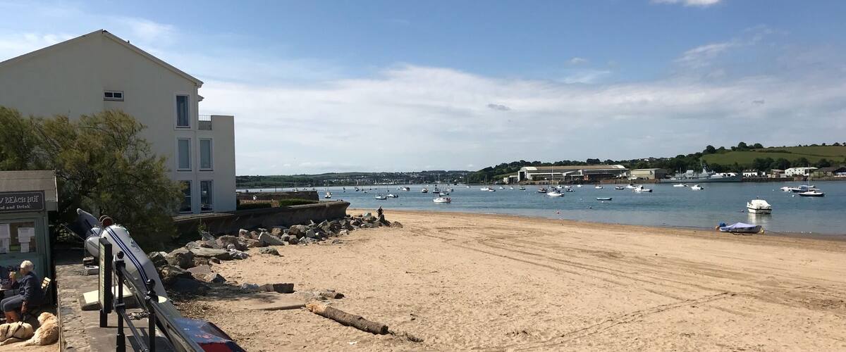 A sandy beach with a white building and a sign that reads "BEACH BAR". Boats are visible on the water in the background.