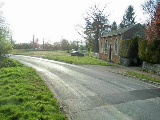 A rural road with a car parked on the side, surrounded by greenery and a brick building.