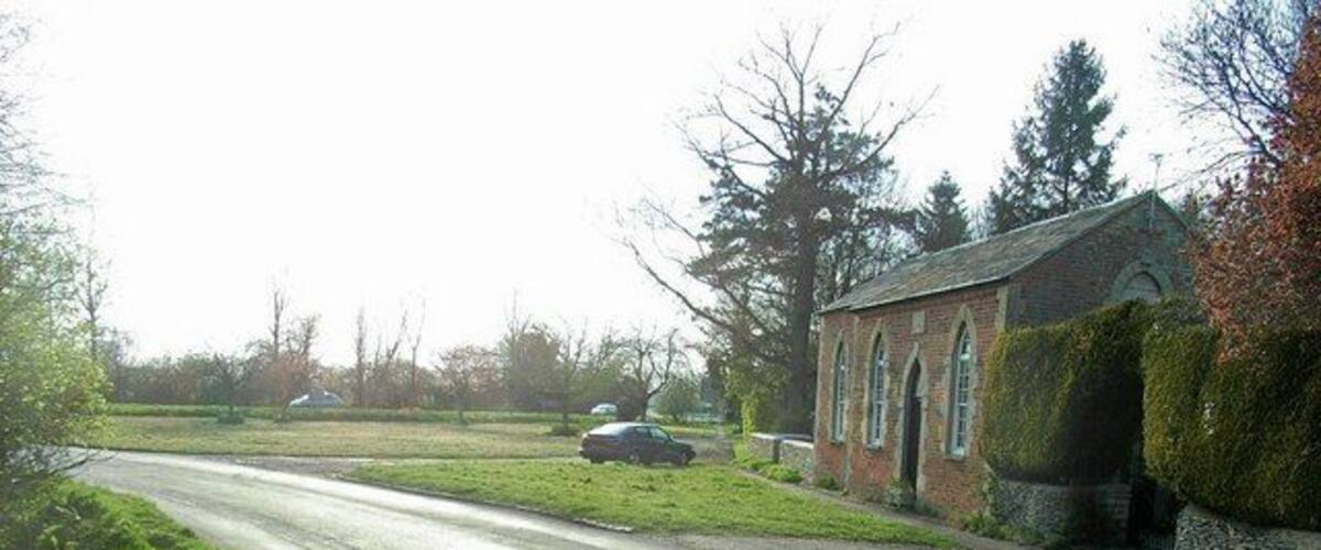 A rural road with a car parked on the side, surrounded by greenery and a brick building.