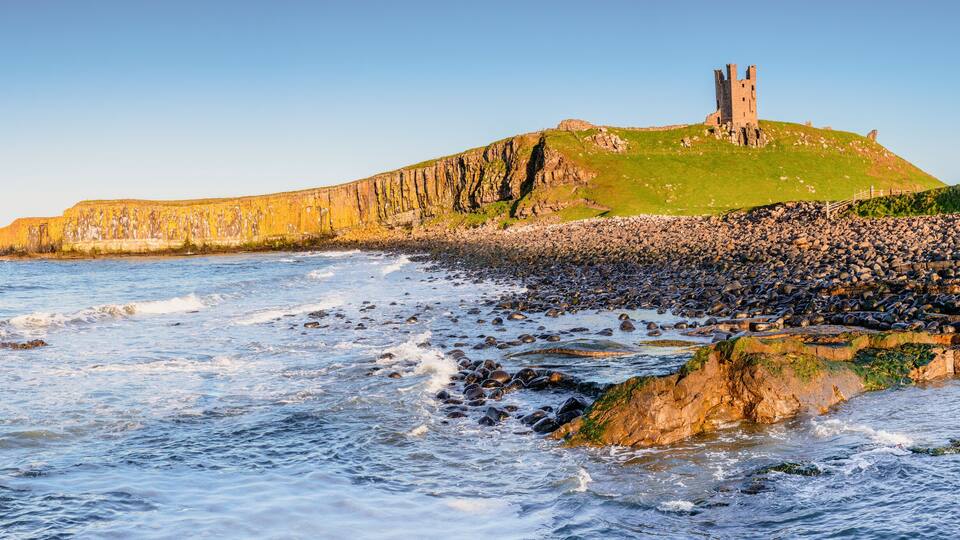 A coastal landscape with a castle on a grassy hill, rocky shoreline, and waves crashing onto the pebbles.