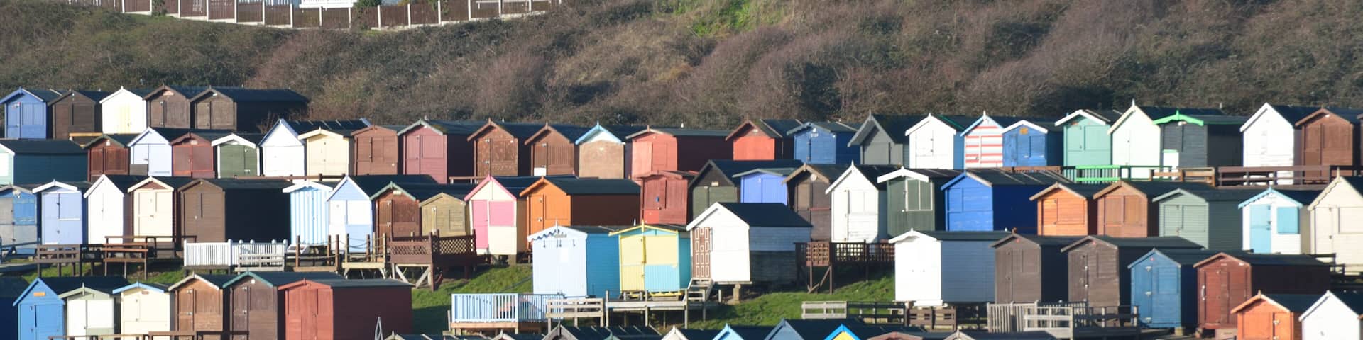 A row of colorful beach huts along a waterfront, with houses in the background on a hillside.