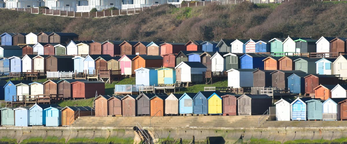A row of colorful beach huts along a waterfront, with houses in the background on a hillside.