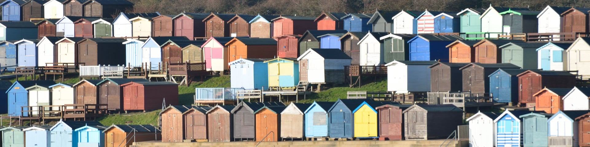 A row of colorful beach huts along a waterfront, with houses in the background on a hillside.