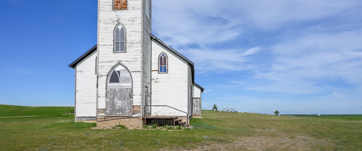 A white church with a cross on top, set in a grassy field under a clear sky.