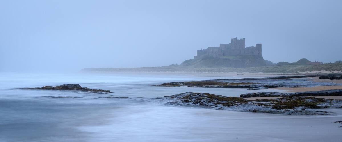 A castle on a cliff overlooking a beach with calm waves.