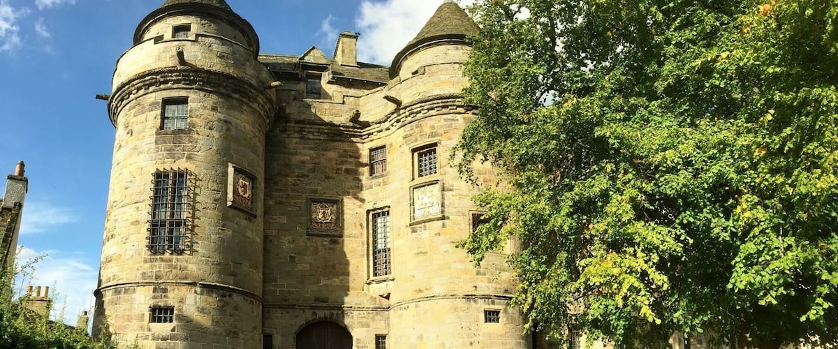 A historic stone castle with two prominent towers, a large arched entrance, and a lush green tree in the foreground.