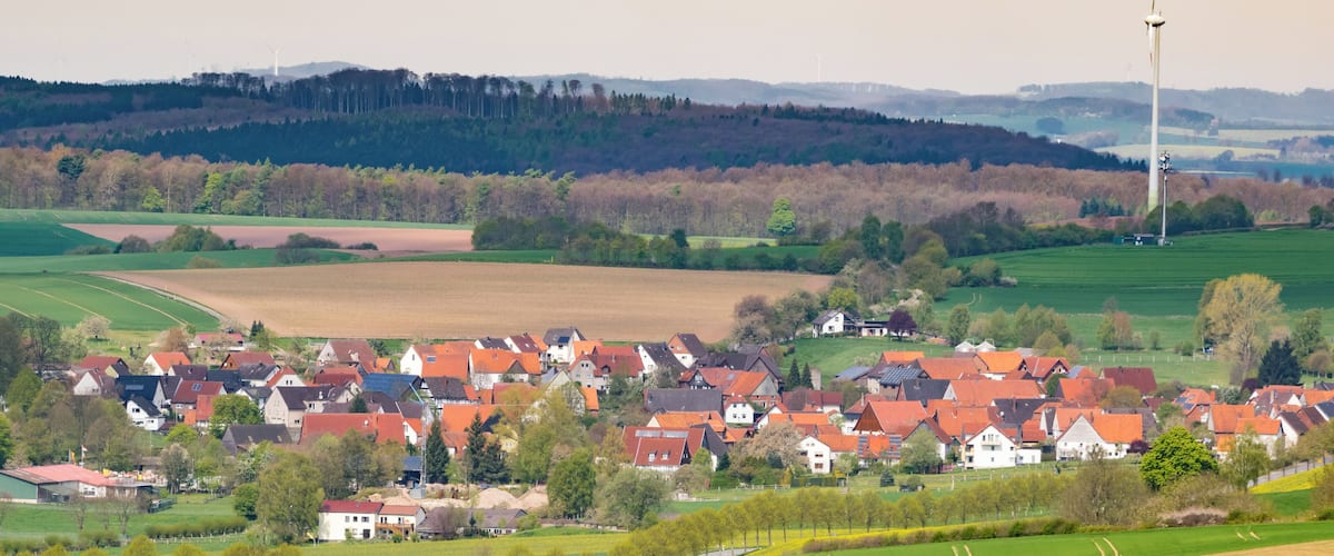 A rural village with red-roofed houses, green fields, and wind turbines in the background.