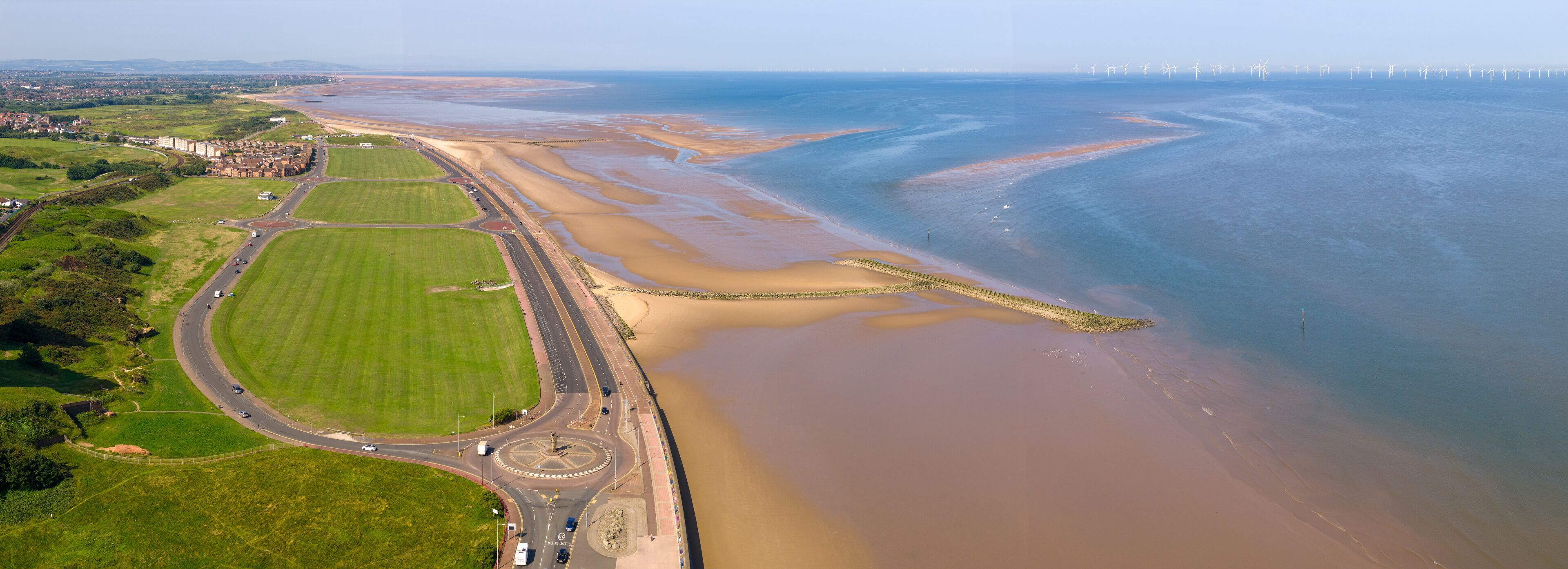 An aerial view of a coastal area with a road, green fields, and a beach with a tidal flat.