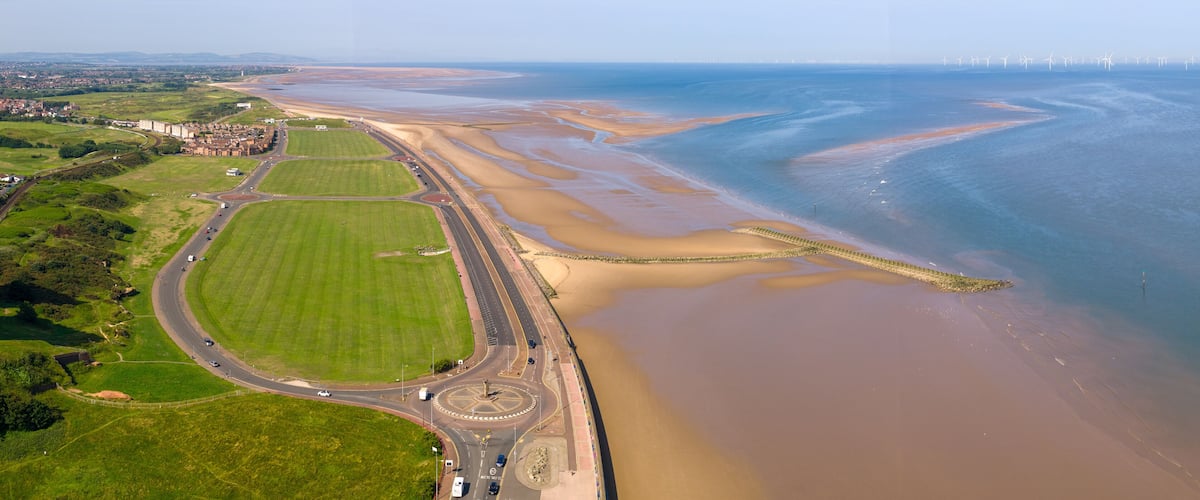 An aerial view of a coastal area with a road, green fields, and a beach with a tidal flat.