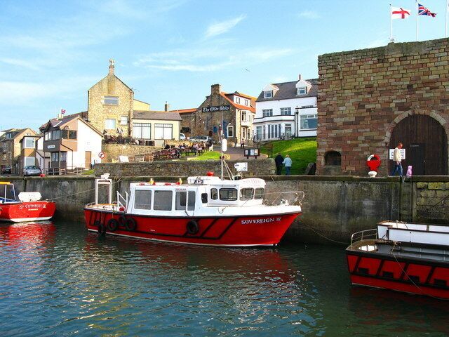 A harbor with red and white boats docked, a stone wall, and buildings in the background.