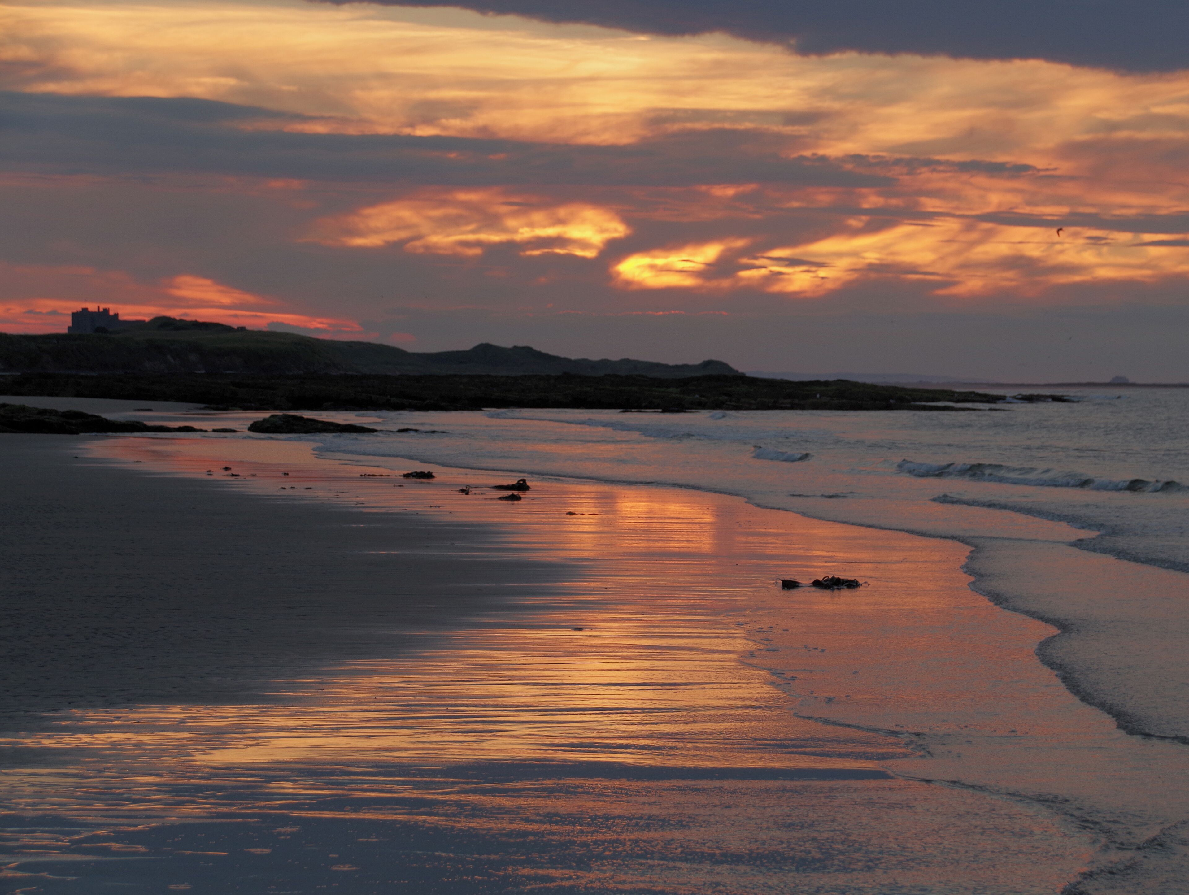 A beach at sunset with waves lapping the shore, a distant castle, and a cloudy sky.