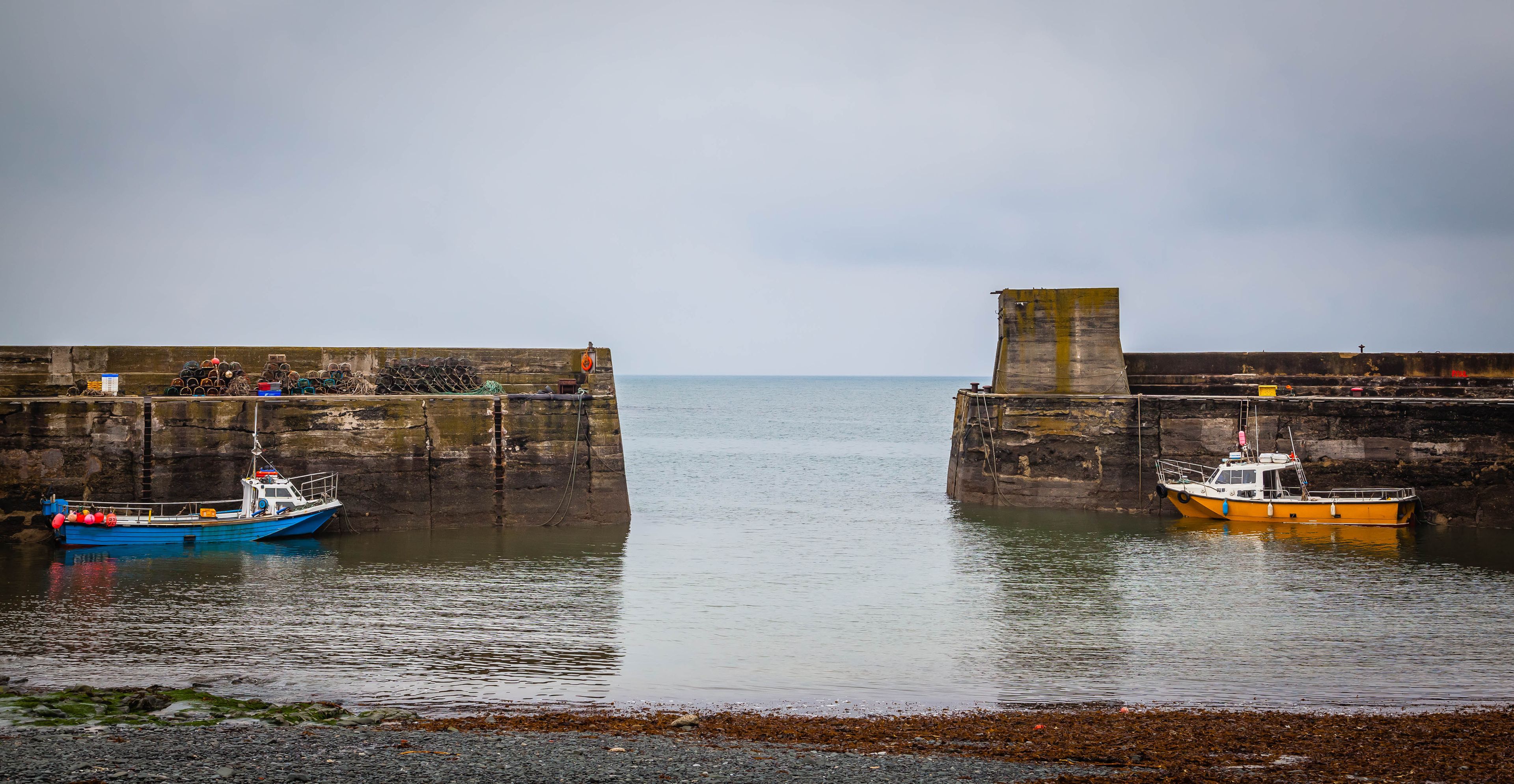 A harbor with two boats docked at a stone pier, calm water, and a cloudy sky.